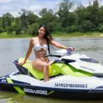 Smiling woman in bikini enjoying a sunny ride on a Sea-Doo jet ski surrounded by lush greenery.